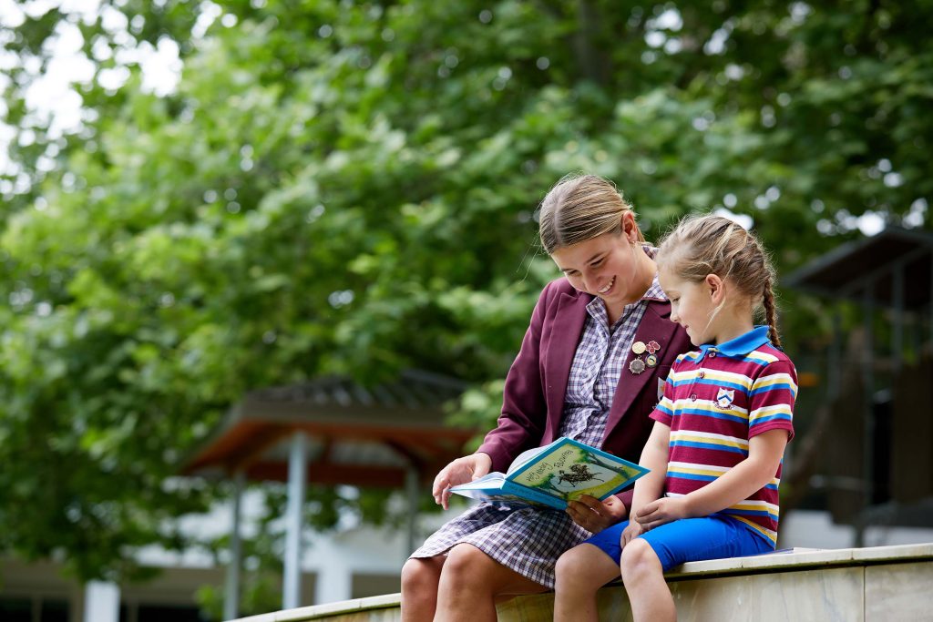 A teenager and child reading book on bench at St Mary's Anglican Girls School.
