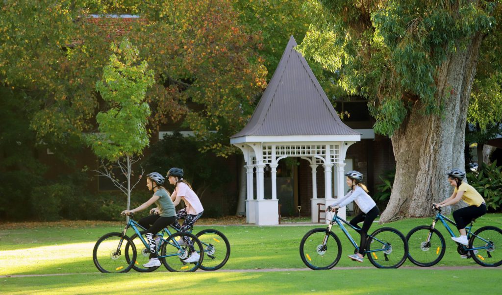 Four people riding bikes in a park at St Mary's Anglican Girls School, surrounded by 16 hectares of green grounds in Karrinyup and Trigg Bushland Reserve. Boarding School In Perth. 