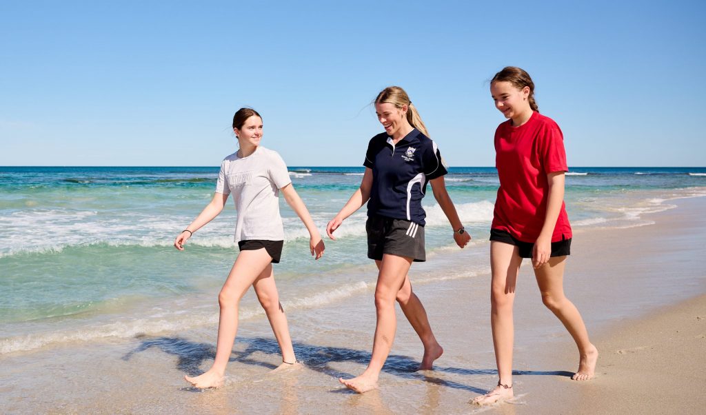 Three young women from St Mary's Anglican Girls School walking on the beach, holding hands. Boarding School In Perth. 